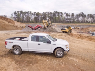 A white pickup truck is parked on a dirt construction site with machinery in the background, including an excavator and a dump truck. The area is surrounded by mounds of dirt and gravel, with a line of trees visible in the distance under a cloudy sky.
