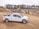 A white pickup truck is parked on a dirt construction site with machinery in the background, including an excavator and a dump truck. The area is surrounded by mounds of dirt and gravel, with a line of trees visible in the distance under a cloudy sky.