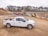 A white pickup truck is parked on a dirt construction site with machinery in the background, including an excavator and a dump truck. The area is surrounded by mounds of dirt and gravel, with a line of trees visible in the distance under a cloudy sky.