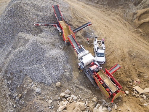 A large industrial machine, likely a rock crusher or screener, is operating in a quarry or construction site. It is surrounded by piles of gravel and rocks. Nearby, there are a couple of white utility vehicles parked on the dirt ground.