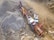 A large industrial machine, likely a rock crusher or screener, is operating in a quarry or construction site. It is surrounded by piles of gravel and rocks. Nearby, there are a couple of white utility vehicles parked on the dirt ground.