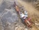 A large industrial machine, likely a rock crusher or screener, is operating in a quarry or construction site. It is surrounded by piles of gravel and rocks. Nearby, there are a couple of white utility vehicles parked on the dirt ground.