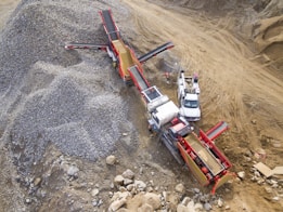 A large industrial machine, likely a rock crusher or screener, is operating in a quarry or construction site. It is surrounded by piles of gravel and rocks. Nearby, there are a couple of white utility vehicles parked on the dirt ground.