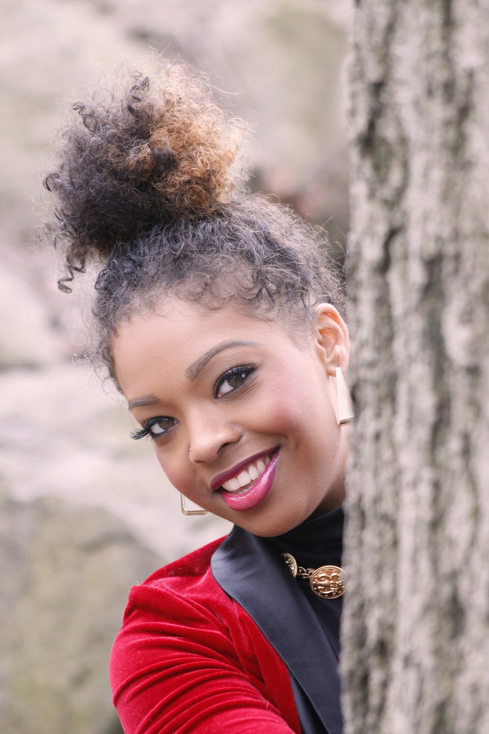 woman wearing black and red blazer hiding behind tall tree while smiling during daytime