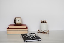 A minimalist workspace setup featuring a stack of three books with a vintage alarm clock on top, a white jar filled with colored pencils, a pair of tortoiseshell glasses, and a closed notebook with earphones resting on top.
