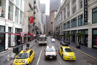 A busy urban street flanked by tall buildings with storefronts, including a store with the word 'REPUBLIC' prominently displayed. Several taxis in bright yellow and other vehicles move along the street. People can be seen walking on the sidewalks, and there's a public bus traveling in the opposite direction.