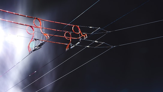 A set of overhead electric wires with red and black insulators against a dark, indistinct background. The lighting creates a contrast between the brightness of the wires and the dark surroundings. There is a noticeable glow on the left side, possibly from a light source.