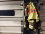 A high-visibility firefighter jacket is hung on a metallic garage door. The jacket features neon yellow and reflective orange stripes, and the word 'ISON' is visible on the back. The setting suggests an industrial or emergency service environment.