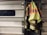 A high-visibility firefighter jacket is hung on a metallic garage door. The jacket features neon yellow and reflective orange stripes, and the word 'ISON' is visible on the back. The setting suggests an industrial or emergency service environment.