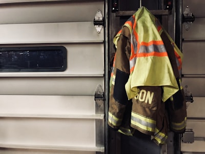 A high-visibility firefighter jacket is hung on a metallic garage door. The jacket features neon yellow and reflective orange stripes, and the word 'ISON' is visible on the back. The setting suggests an industrial or emergency service environment.