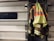 A high-visibility firefighter jacket is hung on a metallic garage door. The jacket features neon yellow and reflective orange stripes, and the word 'ISON' is visible on the back. The setting suggests an industrial or emergency service environment.