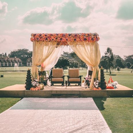 A beautifully decorated outdoor stage featuring two chairs under a canopy draped with sheer yellow curtains adorned with a colorful floral arrangement on top. The setting is in an open grassy area, possibly a park or garden, with trees in the background and a path leading to the stage. There are decorative elements like flower vases and fabric drapes around the stage.