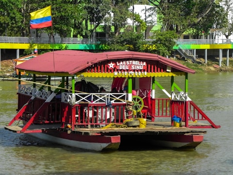 A colorful riverboat with a red and green structure, featuring a roof and seating areas, is docked on a calm waterway. The boat displays a sign with the name 'La Estrella del Sinu' and a date. A Colombian flag flies prominently above the boat. Trees and a building with similar vibrant colors line the riverbank in the background.