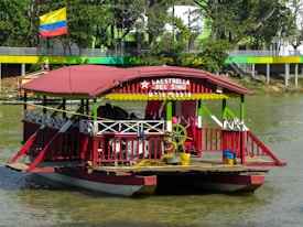 A colorful riverboat with a red and green structure, featuring a roof and seating areas, is docked on a calm waterway. The boat displays a sign with the name 'La Estrella del Sinu' and a date. A Colombian flag flies prominently above the boat. Trees and a building with similar vibrant colors line the riverbank in the background.