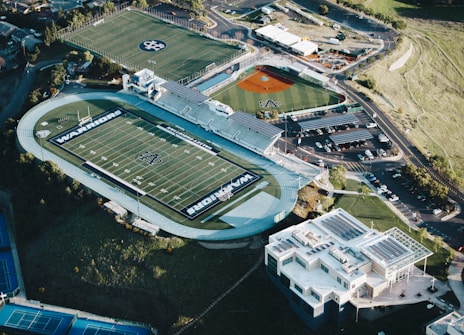 An aerial view of a sports complex featuring a large football stadium with the word 'WARRIOR' painted on the field. Adjacent are fields for other sports, including a baseball diamond and tennis courts. Surrounding the complex are several buildings and parking lots, with nearby roads and green grassy areas visible.