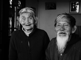 An elderly couple is captured in a candid moment, with both smiling warmly. The image is in black and white, highlighting their expressive faces and the detailed texture of their skin. The background shows an interior setting with framed photographs on the wall.