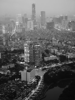 A sprawling cityscape with numerous high-rise buildings reaching into a hazy sky. Streets are lined with countless lights illuminating the densely packed urban environment. A river reflects the glow of the city, winding its way through the metropolis.