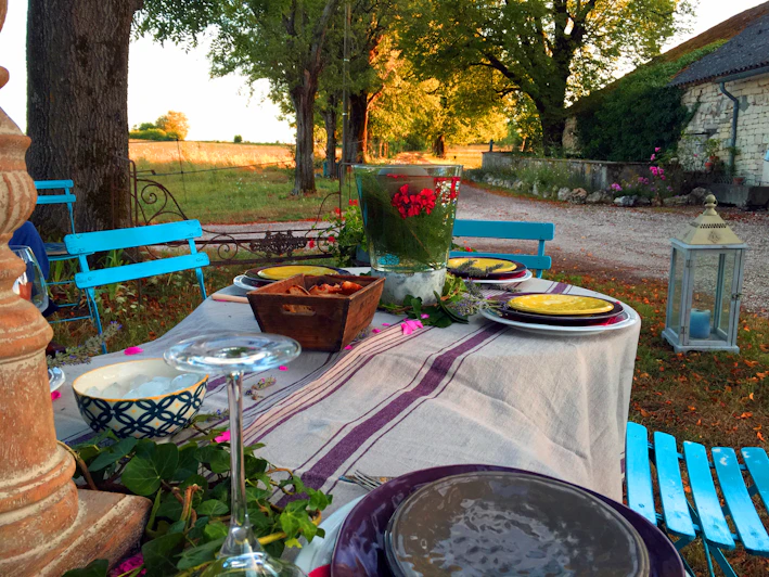 A beautifully set outdoor dining table with a white tablecloth and purple stripes, surrounded by vibrant blue chairs. The table is adorned with dishes, a wooden bowl of food, and a large vase containing bright red flowers. In the background, a tranquil rural landscape features trees and a stone building, adding to the serene atmosphere.