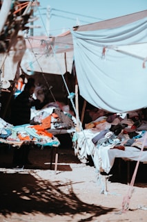 A market scene with tables covered in clothes under a makeshift canopy. Sunlight filters through, casting shadows on the ground. The setting appears rustic and informal, suggesting an outdoor or flea market environment.