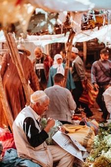 A bustling market scene with people engaged in various activities. An elderly man in the foreground is holding a bunch of greenery and reading a newspaper. He is surrounded by colorful textiles and market goods. Several other people, including a woman in a headscarf, are in the background engaged in conversation and shopping. The setting is warmly lit, with natural sunlight filtering through a makeshift overhead cover made of fabric and sticks.
