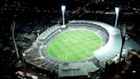 An aerial view of a large, illuminated sports stadium filled with spectators. The stadium is oval-shaped, featuring a green field in the center with markings for a sports game, likely football or rugby. Bright floodlights illuminate the scene, creating a vibrant atmosphere. Surrounding the stadium are urban buildings and streets, barely visible in the night.
