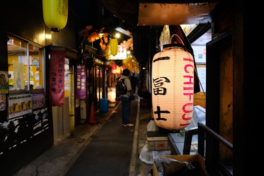 A narrow, dimly lit alley lined with traditional Japanese lanterns and small storefronts. The atmosphere is cozy and inviting, with warm light glowing from the windows. Vibrant signs and posters in Japanese script decorate the walls, and a person with a backpack walks down the path.