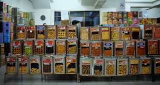 A variety of snacks are displayed in large transparent containers stacked on metal shelves. The containers hold different types of cookies, biscuits, and other snack items, each labeled with a colorful tag. The background includes additional stacked containers and a wall clock, suggesting an organized retail space.