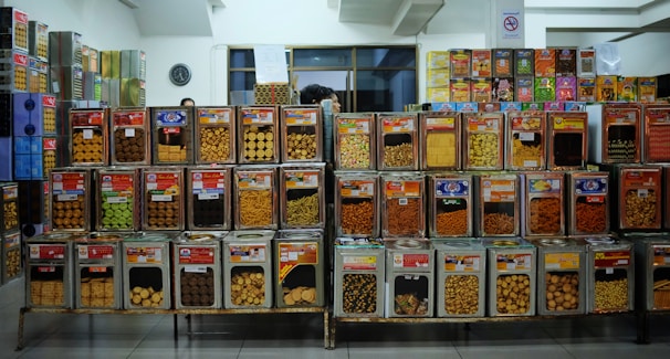 A variety of snacks are displayed in large transparent containers stacked on metal shelves. The containers hold different types of cookies, biscuits, and other snack items, each labeled with a colorful tag. The background includes additional stacked containers and a wall clock, suggesting an organized retail space.