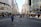 A busy urban street scene with numerous pedestrians walking along a wide road lined with tall commercial buildings. The street appears to be a popular shopping area, with recognizable storefronts and retail signs. People are dressed in a variety of styles, indicating a bustling metropolitan setting. The scene captures the essence of city life with both locals and visitors strolling and interacting.