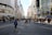 A busy urban street scene with numerous pedestrians walking along a wide road lined with tall commercial buildings. The street appears to be a popular shopping area, with recognizable storefronts and retail signs. People are dressed in a variety of styles, indicating a bustling metropolitan setting. The scene captures the essence of city life with both locals and visitors strolling and interacting.