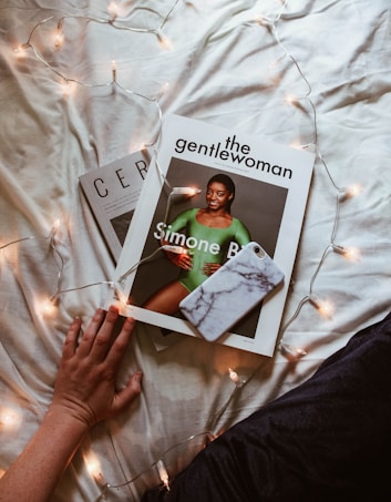 An image depicts a hand resting near a magazine titled 'The Gentlewoman' with a cover featuring a person in a green garment. The magazine is placed on top of another magazine with partially visible text. A smartphone with a marble-like case is resting on the magazine. String lights are artfully draped around, providing a soft glow on the beige fabric surface.
