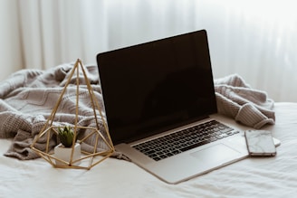 A laptop with a dark screen rests on a bed covered with a textured grey blanket. To the left of the laptop is a decorative item including a small plant in a geometric gold holder. On the right side of the laptop, a smartphone with a marble-like case lies on the bed. Soft light filters through light curtains in the background, creating a cozy and serene atmosphere.