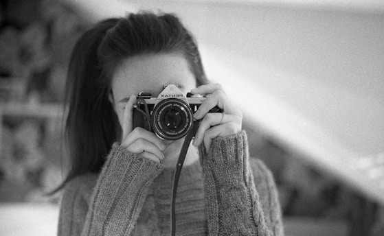 A person holding a vintage camera close to their face, wearing a cozy sweater. The image is in black and white, emphasizing the classic and timeless feel. The focus is on the camera with blurred background.