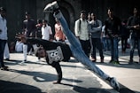 A group of people are gathered in an open area with a young man in the foreground performing a dynamic breakdancing move, balancing on one hand with one leg extended upward. The spectators are casually dressed, watching the performance, creating an engaging street scene.