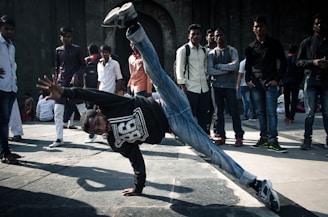 A group of people are gathered in an open area with a young man in the foreground performing a dynamic breakdancing move, balancing on one hand with one leg extended upward. The spectators are casually dressed, watching the performance, creating an engaging street scene.