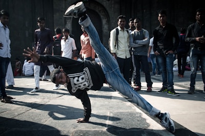 A group of people are gathered in an open area with a young man in the foreground performing a dynamic breakdancing move, balancing on one hand with one leg extended upward. The spectators are casually dressed, watching the performance, creating an engaging street scene.