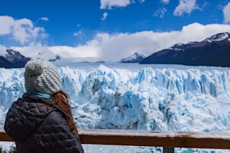A person dressed in a winter coat and a knit hat with a pompom is looking at a large glacier. The glacier is expansive with jagged ice formations, set against a backdrop of snow-capped mountains and a bright, partly cloudy sky.