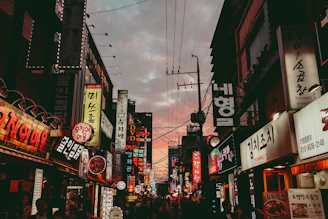 A vibrant urban street corner at dusk with glowing orange and red neon signs reflecting the energy of a lively community.