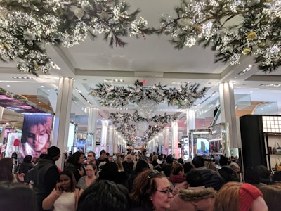 A crowded shopping mall adorned with festive holiday decorations, including garlands and large ornaments. Numerous people are shopping and walking through the main aisle. The setting is bright and bustling with a lively atmosphere.