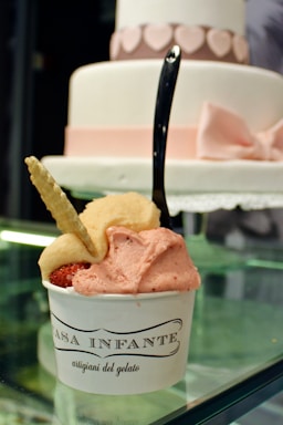 A cup of gelato with strawberry and vanilla flavors is garnished with a wafer and a cookie. In the background, there is a tiered cake decorated with hearts and a pink bow.
