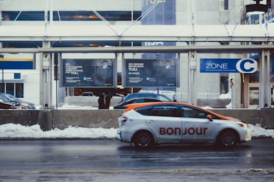A city street scene with a vehicle marked 'Bonjour' in the foreground, traveling near a snow-covered pavement. Behind the car, there is a series of advertisements on a concrete barrier and the street appears wet. The background features a bus stop sign labeled 'Zone C' and a modern building, suggesting a busy urban environment.