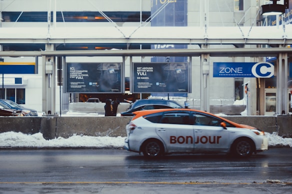 A city street scene with a vehicle marked 'Bonjour' in the foreground, traveling near a snow-covered pavement. Behind the car, there is a series of advertisements on a concrete barrier and the street appears wet. The background features a bus stop sign labeled 'Zone C' and a modern building, suggesting a busy urban environment.