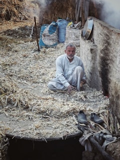 An elderly man sits on a surface covered with straw and husks, surrounded by a rustic environment with bags labeled 'Not for Sale' and traditional tools. He appears contemplative, with various household items and signs of agriculture in the background.