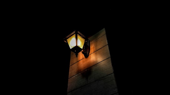 A single illuminated wall-mounted lantern casts a warm glow on a building wall, surrounded by darkness.
