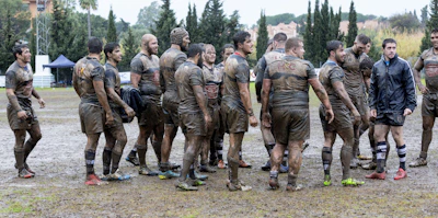 A group of coaches discussing strategies with clipboards beside a rugby pitch.