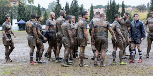 A group of muddy rugby players are gathered on a wet and messy rugby field. They appear to be discussing the game or taking a break, with some wearing shorts and others in longer attire. The background features trees and a couple of tents.