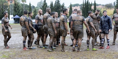 A group of muddy rugby players are gathered on a wet and messy rugby field. They appear to be discussing the game or taking a break, with some wearing shorts and others in longer attire. The background features trees and a couple of tents.