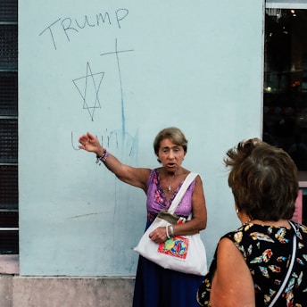 An older woman wearing a purple sleeveless top and holding a white bag is gesturing towards a wall with graffiti. The graffiti includes the word 'TRUMP', a Star of David, a cross, and another word that's partially visible. Another woman with short hair listens, wearing a dark floral-patterned top.