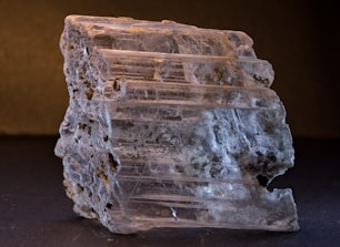 A close-up of a rough, translucent mineral specimen with visible striations and textured surfaces. The mineral displays varying shades of gray and white, with some darker inclusions. The lighting highlights the layered structure, emphasizing the natural beauty of the mineral.