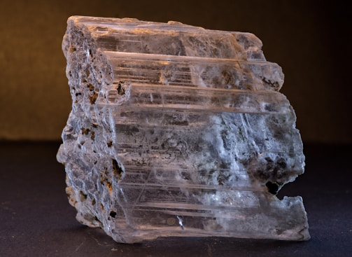 A close-up of a rough, translucent mineral specimen with visible striations and textured surfaces. The mineral displays varying shades of gray and white, with some darker inclusions. The lighting highlights the layered structure, emphasizing the natural beauty of the mineral.